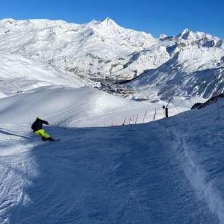 Skier turning on the Merles red piste below the Merles chairlift, with Tignes Le Lac and snowy mountains in the distance.