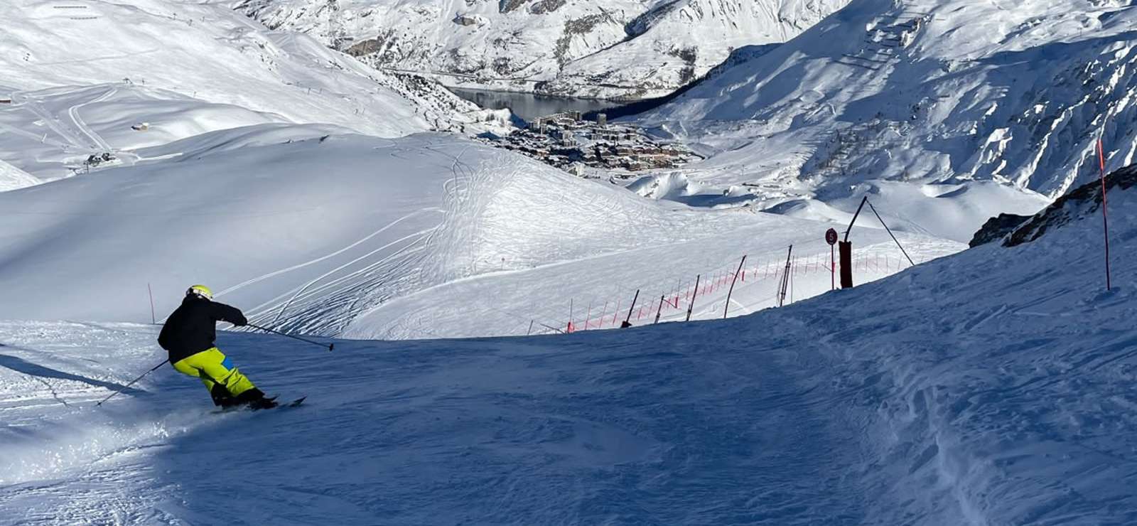 Skier turning on the Merles red piste below the Merles chairlift, with Tignes Le Lac and snowy mountains in the distance.