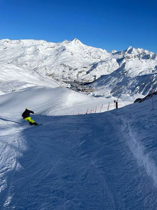 Skier turning on the Merles red piste below the Merles chairlift, with Tignes Le Lac and snowy mountains in the distance.