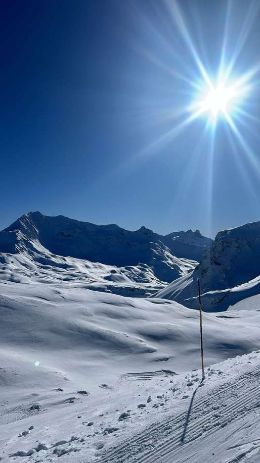 Sunlit snow-covered peaks beside the Génépy blue piste in Tignes, with groomed snow and a piste marker in the foreground.